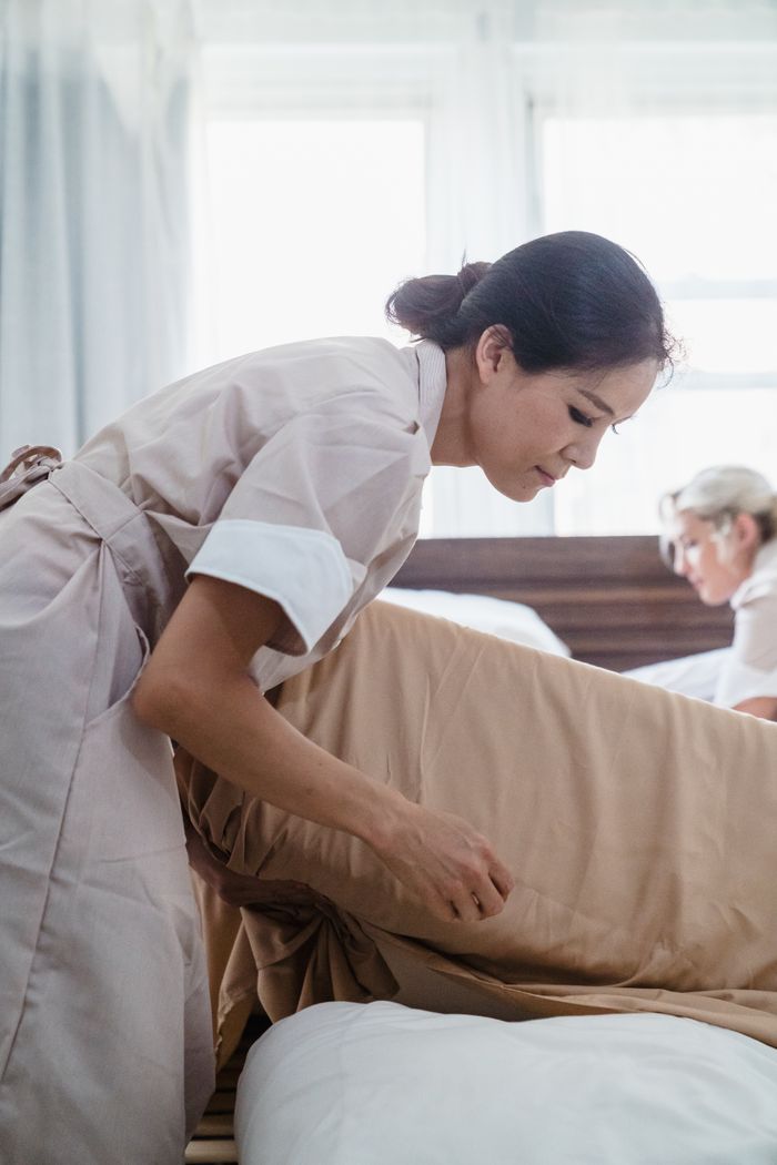 our-story Hotel staff in uniform making a bed with precision and care in a bright room setting.