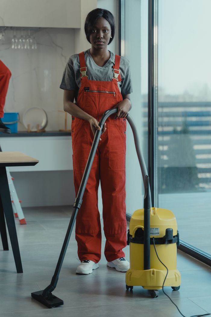 gallery-04 An adult woman in red coveralls operates a vacuum cleaner in a modern indoor setting, focusing on cleaning tasks.